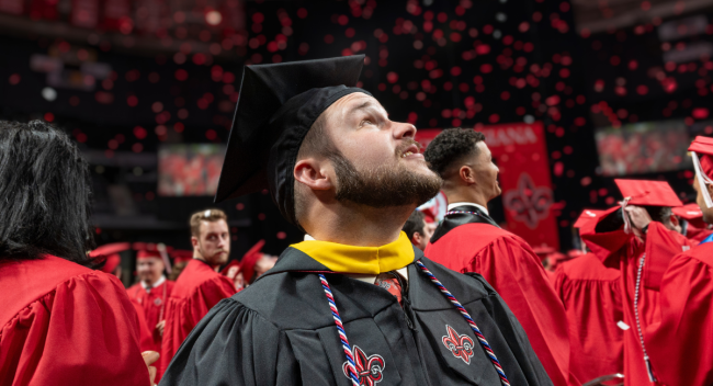 Online master's graduate wears a black cap and gown, gazing up as red confetti falls.